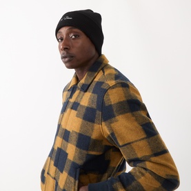 Model with Les Deux Signature Logo Beanie Hat in black ribbed knit with embroidered logo, styled with yellow and navy checked overshirt.