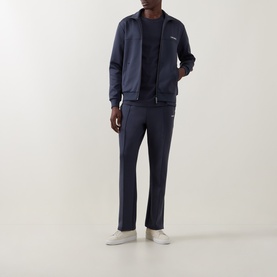 Model wearing Les Deux India Ink Blue sweatpants in polyester-elastane blend, styled with matching zip-through jacket and white trainers.