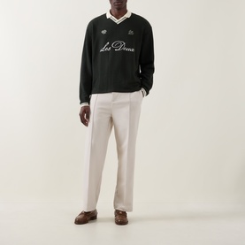 Model wearing Les Deux Rosin Green sweatshirt with V-neck and script logo, styled with cream wide-leg trousers and brown loafers