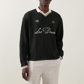 Model wearing Les Deux rosin green ribbed cotton sweatshirt with contrast V-neck collar and embroidered script logo, styled with cream trousers