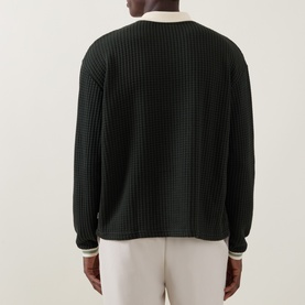 Model wearing Les Deux rosin green waffle-knit sweatshirt, back view, with cream contrast collar and cuff trims, styled with white trousers.