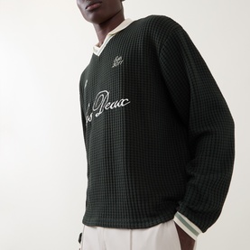 Model wearing Les Deux Rosin Green waffle-knit sweatshirt with cream V-neck trim and script logo embroidery on chest.