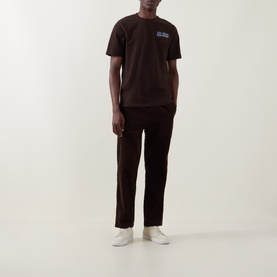 Model wearing Les Deux Record Logo Back Graphic T-Shirt in Mole Brown with matching brown trousers and white trainers.