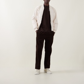 Model wearing Les Deux Record Logo Back Graphic T-Shirt in Mole Brown, styled with cream bomber jacket and dark corduroy trousers.