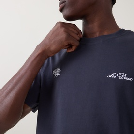 Model wearing Les Deux India Ink Blue cotton T-shirt with white embroidered logo and floral motif on chest, close-up detail shot