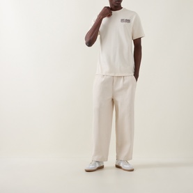 Model wearing Les Deux ivory cotton T-shirt with small chest graphic, styled with matching cream wide-leg trousers and grey trainers.