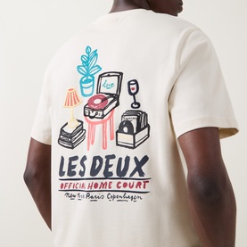 Model wearing Les Deux Ivory cotton T-shirt, back view showing colourful Official Home Court graphic print with record player and lamp illustration.