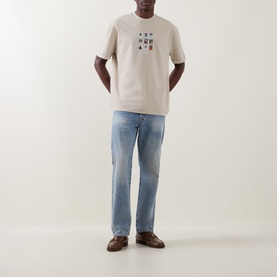 Model wearing Les Deux light sand cotton T-shirt with graphic print, styled with light blue straight-leg jeans and brown loafers.