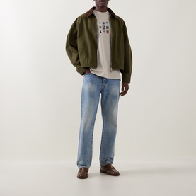 Model wearing Les Deux light sand cotton T-shirt, styled with olive bomber jacket, light wash jeans and brown loafers