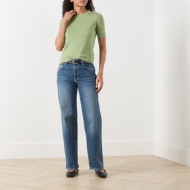 Model wearing Seven Jeans mid blue tapered denim jeans, styled with a green short-sleeve top and black flat shoes.