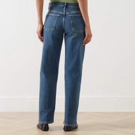 Model wearing Seven Jeans mid blue tapered jeans, rear view showing back pockets and relaxed straight leg, styled with black shoes.