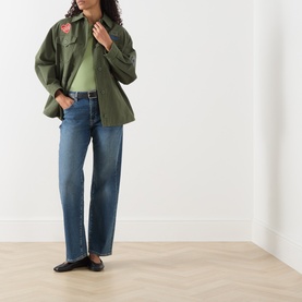 Model wearing Seven Jeans Mid Blue tapered jeans, styled with a khaki military shirt jacket, green top and black loafers.