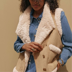 Model wearing Mint Velvet brown shearling gilet with cream fleece lapels, styled with a blue denim shirt and gold-tone jewellery.