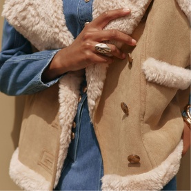 Model wearing Mint Velvet brown shearling-style gilet with cream faux-fur trim, styled over a denim shirt, with a silver statement ring.