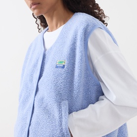 Model wearing American Vintage Hoktown Logo Gilet in Givre Chine blue fleece with logo badge, styled over a white long-sleeve top.