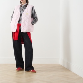 Model wearing American Vintage pink quilted gilet over striped top and red shirt, styled with black wide-leg trousers and red flat shoes.