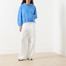 Model wearing American Vintage Plizzy Logo Hoodie in nautique vintage blue with white wide-leg trousers and white trainers.