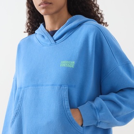 Model wearing American Vintage Plizzy Logo Hoodie in Nautique Vintage blue with neon green logo, pouch pocket and dropped shoulders visible.