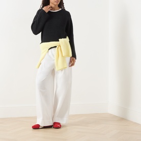 Model wearing AMERICAN VINTAGE Vitow Knitted Sweater in black with white wide-leg trousers, yellow sweater tied at waist, and red flats.