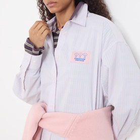 Model wearing American Vintage shirt in Rayures Douceur stripe, featuring chest logo patch, styled with a pink tied sweatshirt and striped top.