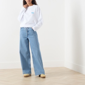 Model wearing American Vintage Gixy Logo Long-Sleeved Top in white with crew neck and chest logo, styled with wide-leg light blue jeans.