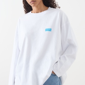 Model wearing American Vintage Gixy Logo Long-Sleeved Top in white with blue chest logo, crew neck, dropped shoulders, styled with blue jeans.
