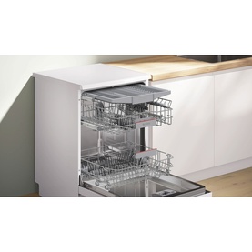 Freestanding dishwasher with open door revealing three wire basket racks, set in a white modern kitchen with wooden worktop.