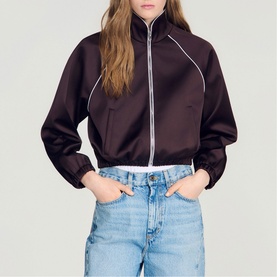 Model wearing Sandro brown cropped zip-up jacket with white piping detail and funnel neck, styled with light blue denim jeans
