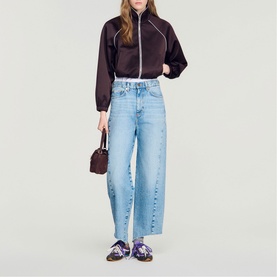Model wearing Sandro brown cropped zip-up jacket with wide-leg light blue jeans and purple loafers, carrying a small brown bag.