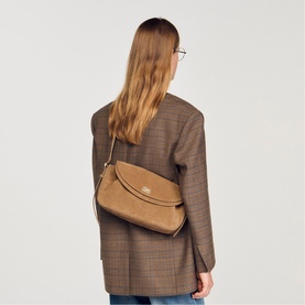 Model carrying Sandro Le Mambo Suede Shoulder Bag in clear, featuring gold-tone hardware and double flap, worn cross-body over a brown blazer.