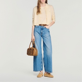 Model carrying a tan structured top-handle bag with stud detailing, styled with cream jacket, cropped top, and wide-leg blue jeans.