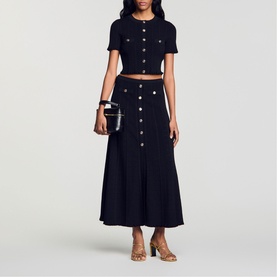 Model wearing Sandro Womens French Dress in black, short-sleeve crop top and midi skirt co-ord with silver buttons, styled with tan sandals.
