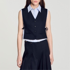Model wearing navy blue tailored waistcoat with button front and side ties, styled with light blue shirt and matching pleated skirt