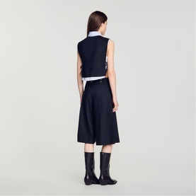 Model wearing navy sleeveless belted waistcoat with white collar shirt, midi skirt, and black knee-high boots, rear view