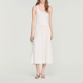 Model wearing Sandro Womens French Dress in clear white, sleeveless knit midi with pleated skirt, styled with silver sandals and clutch bag