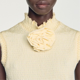 Model wearing pale yellow textured sleeveless high-neck top with oversized ruffle corsage brooch detail at chest