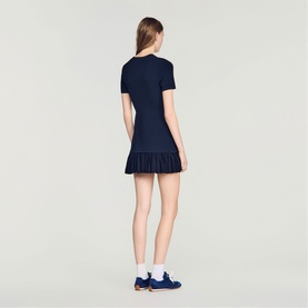 Model wearing Sandro Ruffle Mini Knitted Dress in midnight blue, short sleeves, ruffled hem, styled with white socks and blue shoes.