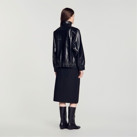 Model wearing Sandro black high-shine leather bomber jacket, styled with black midi skirt and knee-high boots, back view