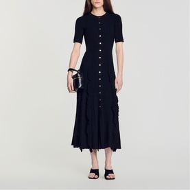 Model wearing Sandro Womens French Dress in black, ribbed knit midi length with button front and ruffle skirt detail, styled with black heeled mules