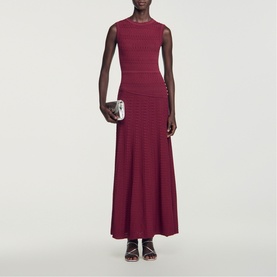 Model wearing Sandro Womens French Dress in red, sleeveless ribbed knit maxi with flared hem, styled with black strappy sandals and white clutch bag.