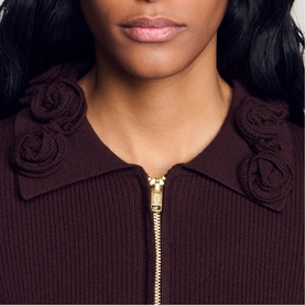 Model wearing Sandro brown ribbed zip-up knit top with 3D rosette collar detail and gold-tone zip, close-up view.