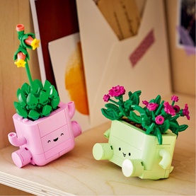 Lego Botanical toy set featuring two smiling watering can plant pots in pink and green with colourful flower builds on a wooden shelf