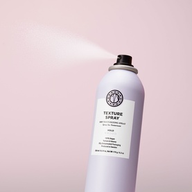 Maria Nila Texture Spray aerosol can in pale grey, shown actively dispensing mist against a pink background