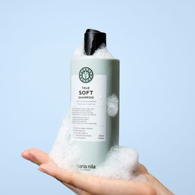 Maria Nila True Soft Shampoo in pale sage green bottle held on a foam-covered hand against a light blue background