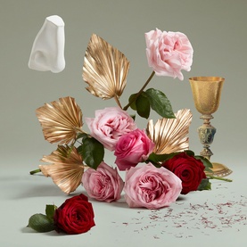 Penhaligon EDP fragrance lifestyle shot with pink and red roses, gold palm leaves, a gold chalice, and a white bottle stopper on a sage green