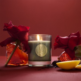 Penhaligon candle in dark glass vessel with gold emblem, lit flame, styled with red roses and lemon slice on deep red background