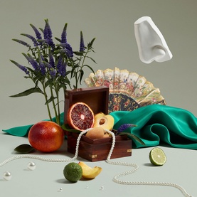 Penhaligon EDP lifestyle shot with pearl necklace, blood oranges, lime, tropical flowers, green silk drape and decorative fan arrangement