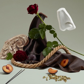 Penhaligon EDP lifestyle shot featuring a white bottle form, dark roses, draped brown fabric, rope basket, and sliced peaches on sage green background