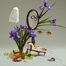 Penhaligon EDP lifestyle shot with purple iris flowers, oval mirror, miniature chaise longue, pearl necklace, and gold-framed glasses on grey
