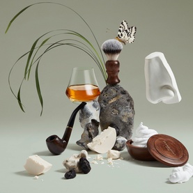 Penhaligon EDP fragrance lifestyle shot featuring whisky glass, shaving brush, pipe, stone, and nose sculpture on sage green background